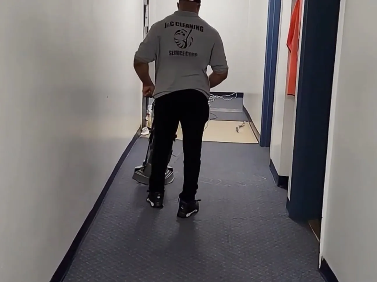 A J&C Cleaning Services technician, seen from behind, running a carpet machine down a carpeted commercial corridor