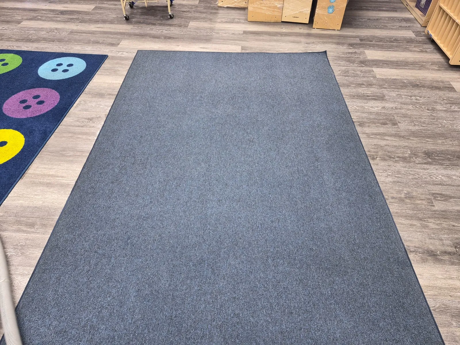 Una alfombra comercial gris recién lavada, tendida sobre el piso de madera de un salón de clases, junto a un tapete infantil estampado