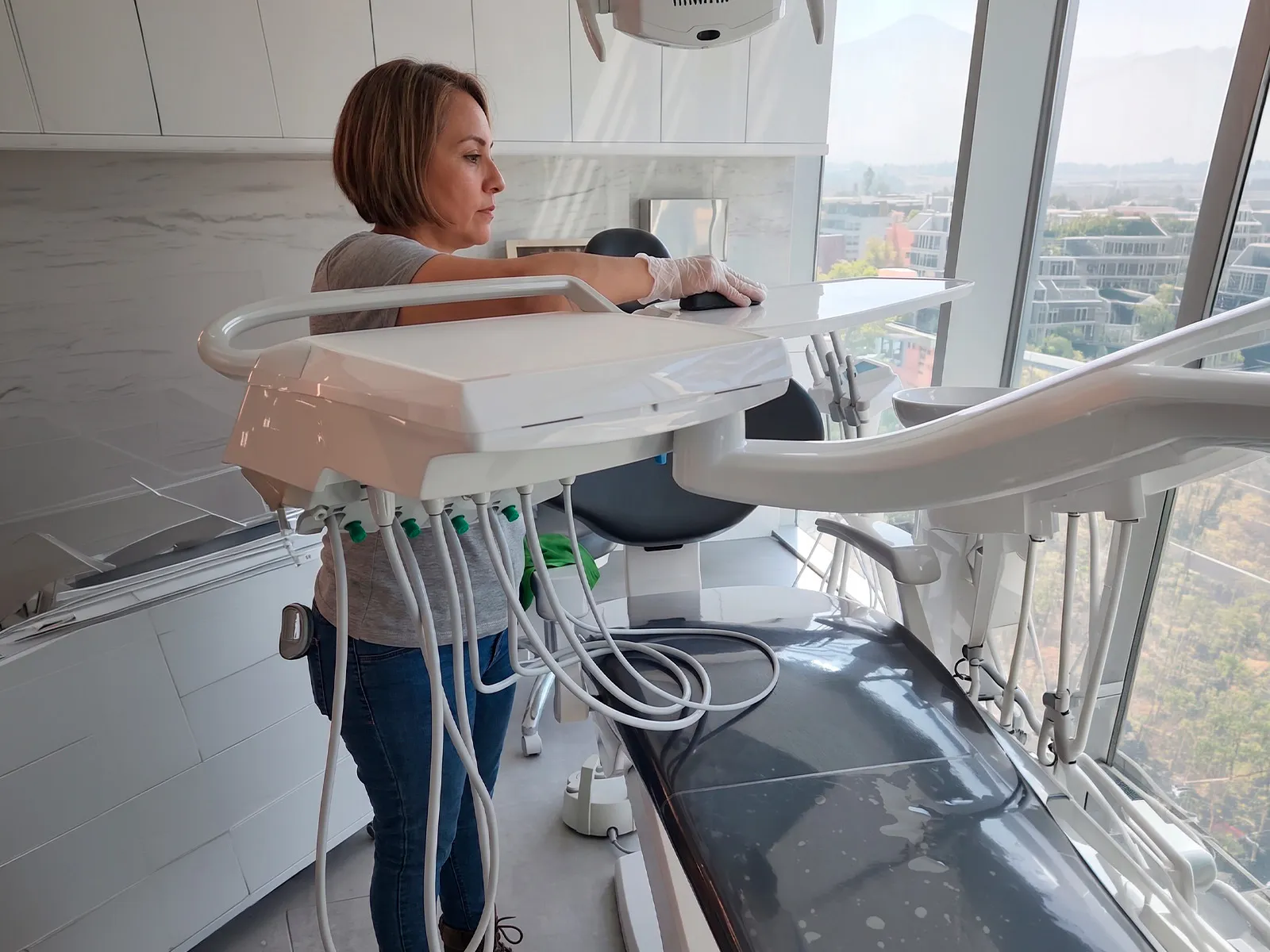 A cleaning technician in a disposable glove wiping down the instrument tray of a dental treatment unit in a daylit clinic