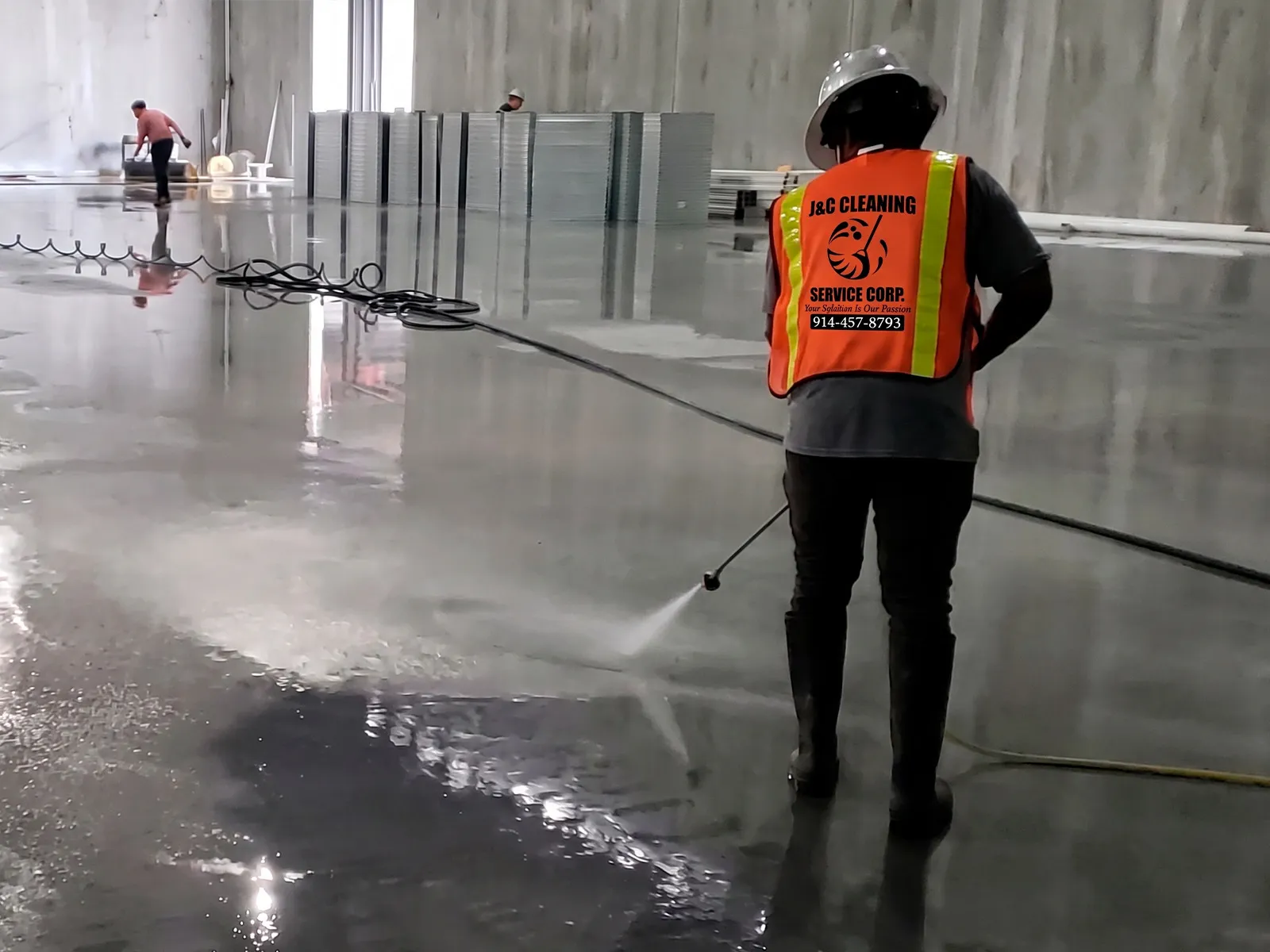 A J&C Cleaning Services worker in a hard hat and high-visibility vest rinsing the concrete floor of a building still under construction