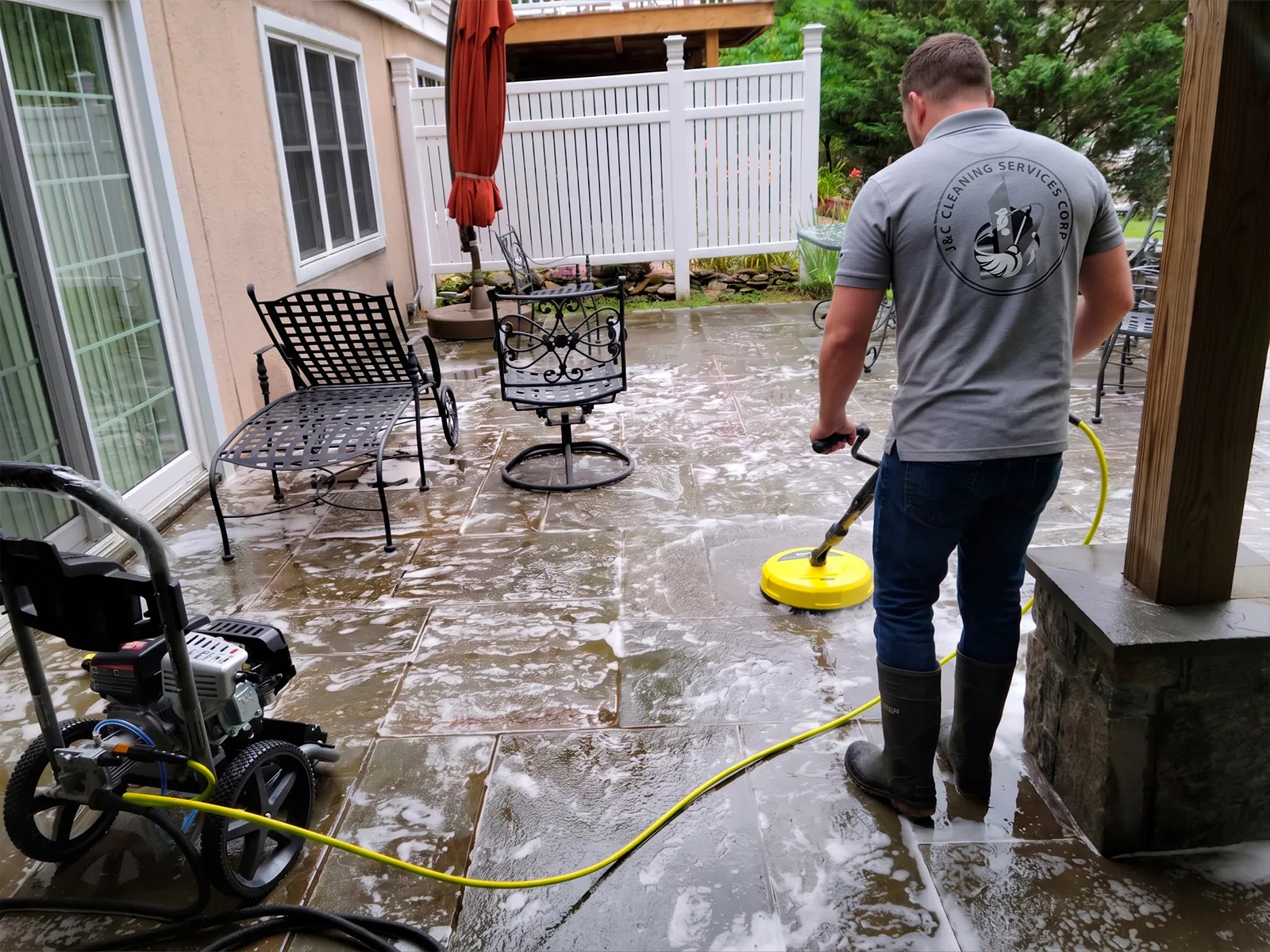 Un técnico de J&C Cleaning Services, con el polo corporativo y botas de trabajo, pasando una limpiadora rotativa de superficies por un patio pavimentado